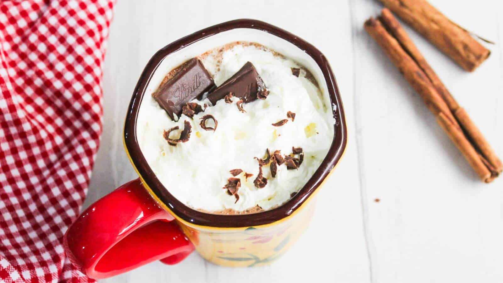 A mug of hot chocolate topped with whipped cream and chocolate shavings, next to a red checkered cloth and cinnamon sticks.