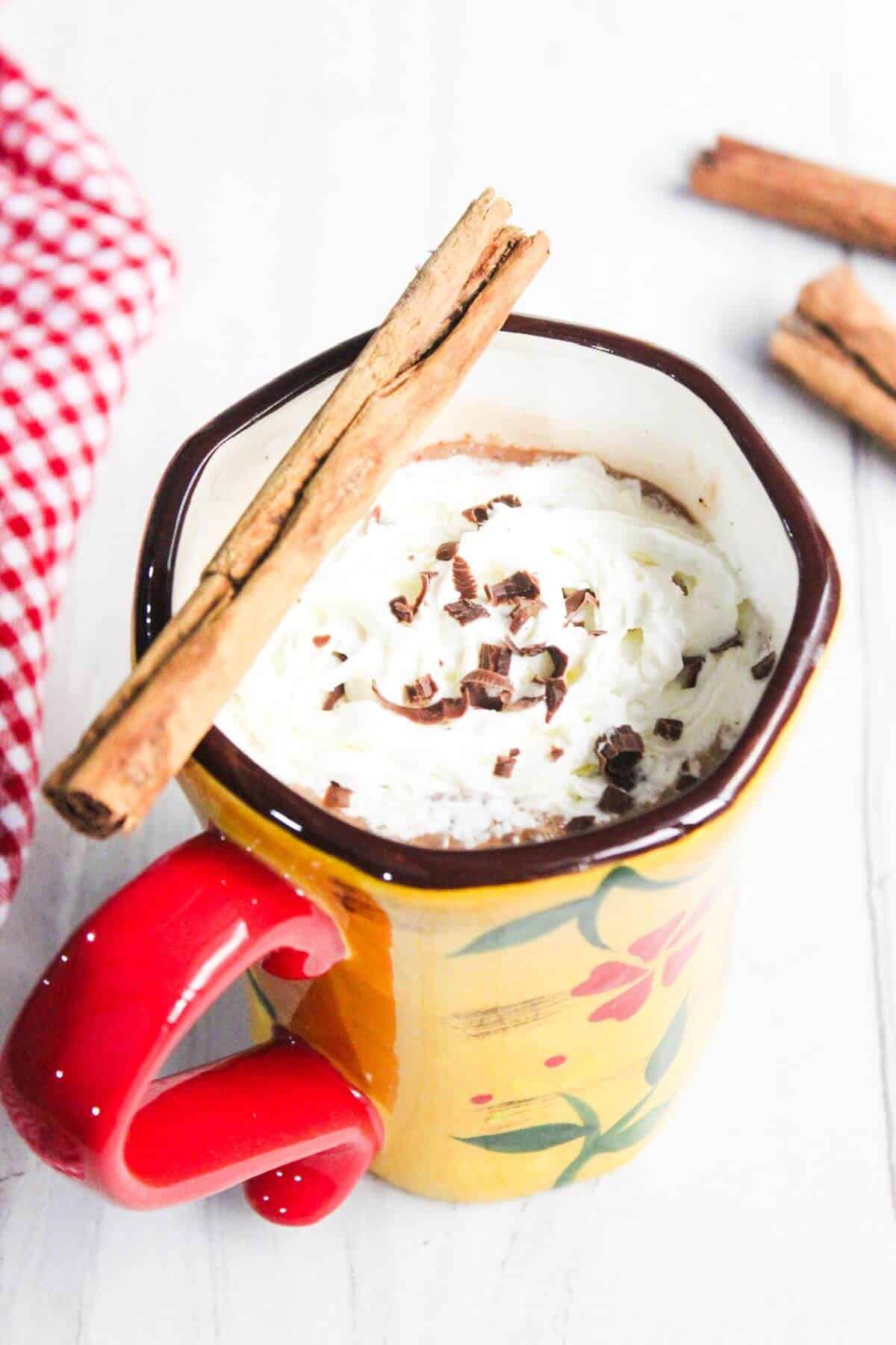 A yellow mug with a red handle, filled with hot chocolate topped with whipped cream, chocolate shavings, and a cinnamon stick. A red and white cloth is beside the mug.