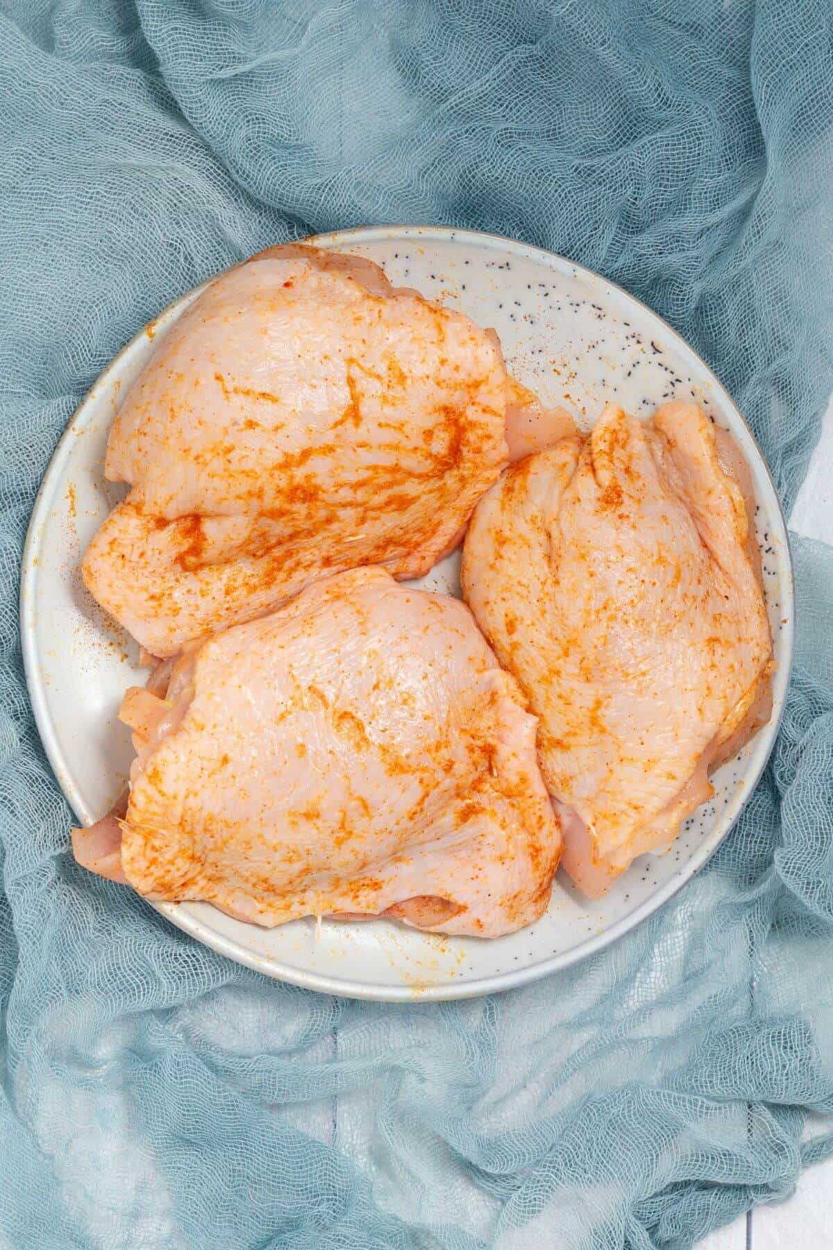 A plate of three raw, seasoned chicken thighs on a blue cloth background.