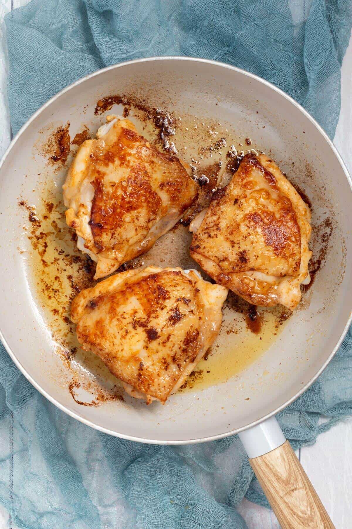 Three cooked chicken thighs with golden brown skin in a frying pan on a teal cloth.