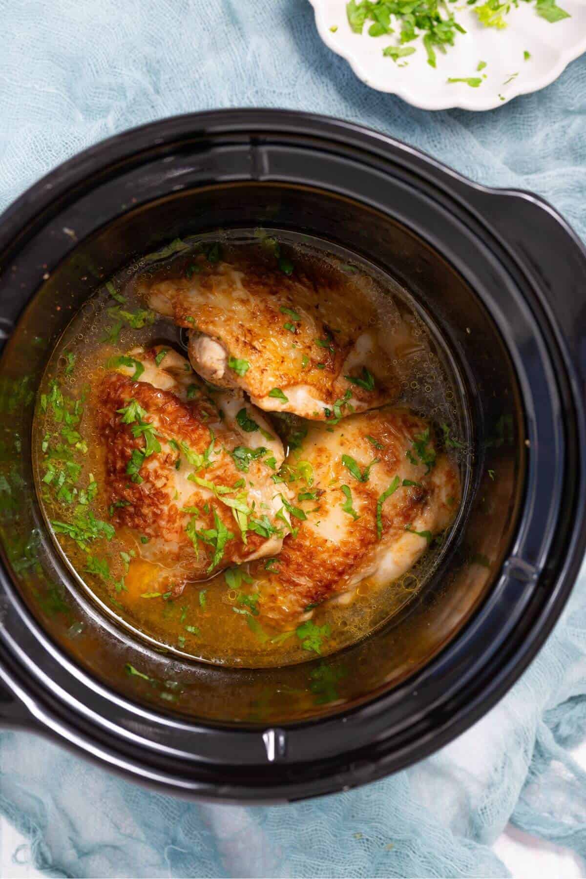 Overhead view of cooked chicken pieces garnished with chopped herbs in a slow cooker filled with broth.