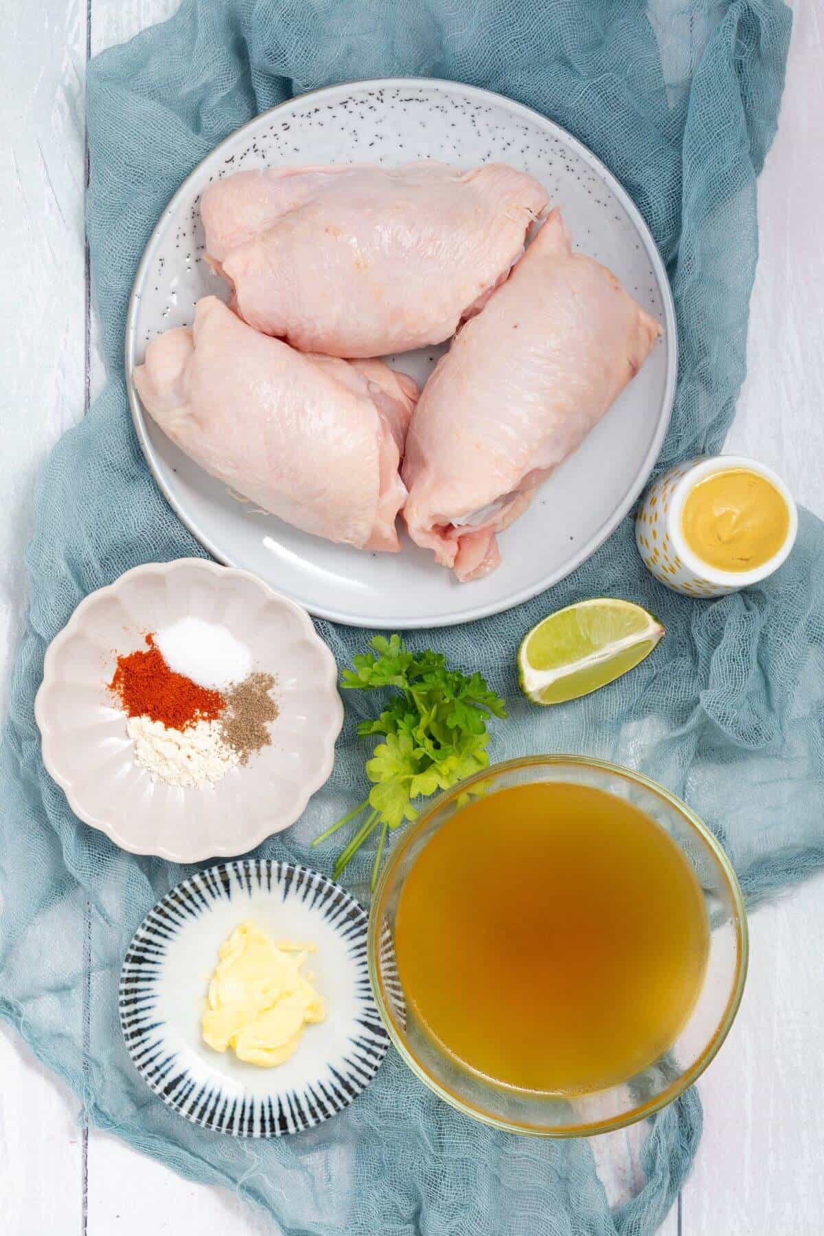 Raw chicken pieces on a plate, surrounded by bowls of spices, butter, a cup of broth, a lemon wedge, and a jar of mustard, all on a blue cloth.