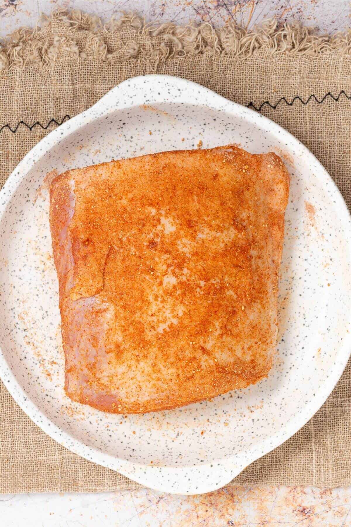 A seasoned raw pork roast in a speckled white dish on a burlap mat.