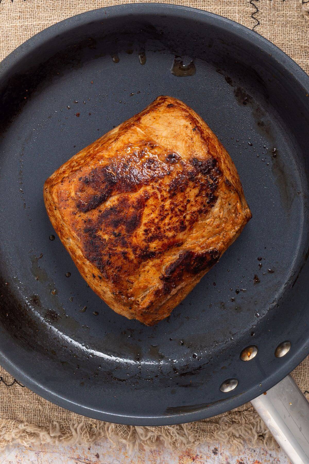 A browned piece of meat in a frying pan on a textured surface.