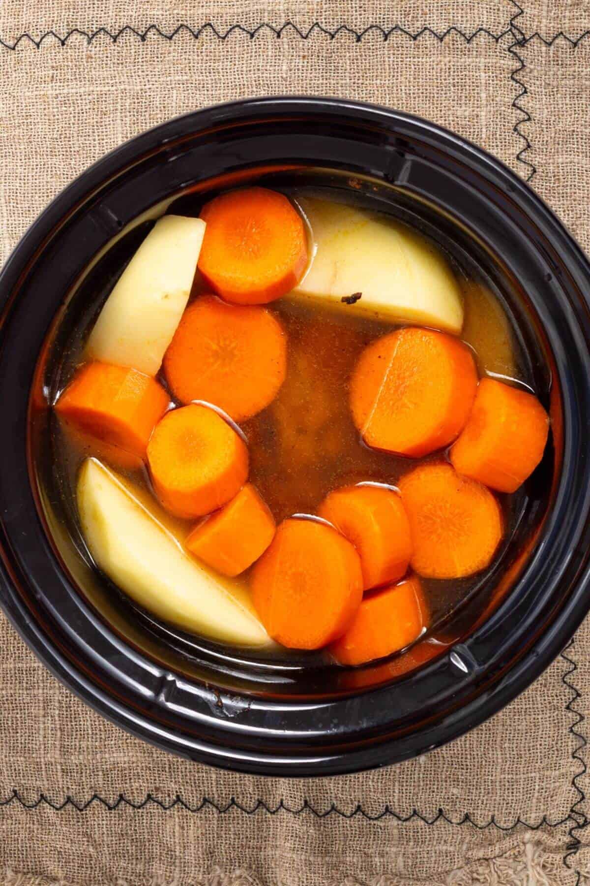 A slow cooker filled with sliced carrots and potato wedges in broth on a textured cloth background.