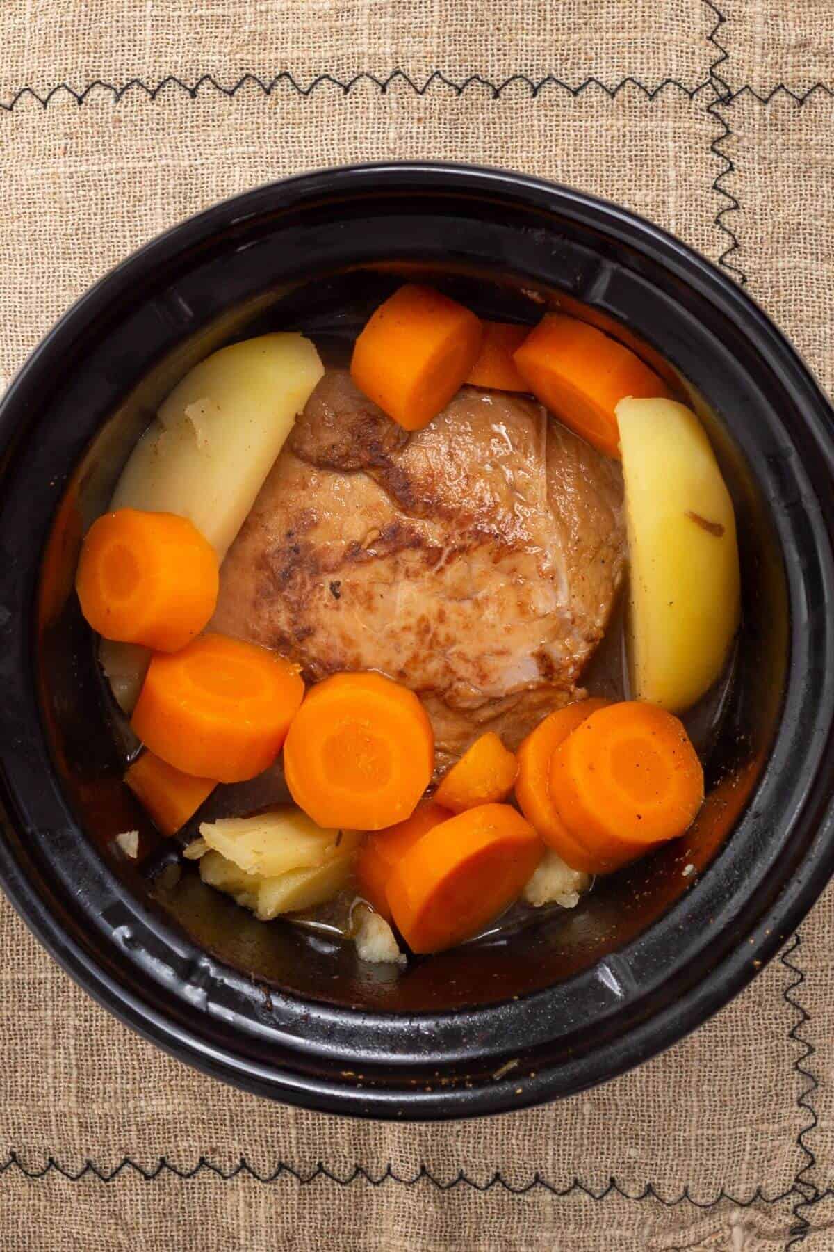 A top view of a slow-cooked pork roast with sliced carrots and potatoes in a black crockpot.