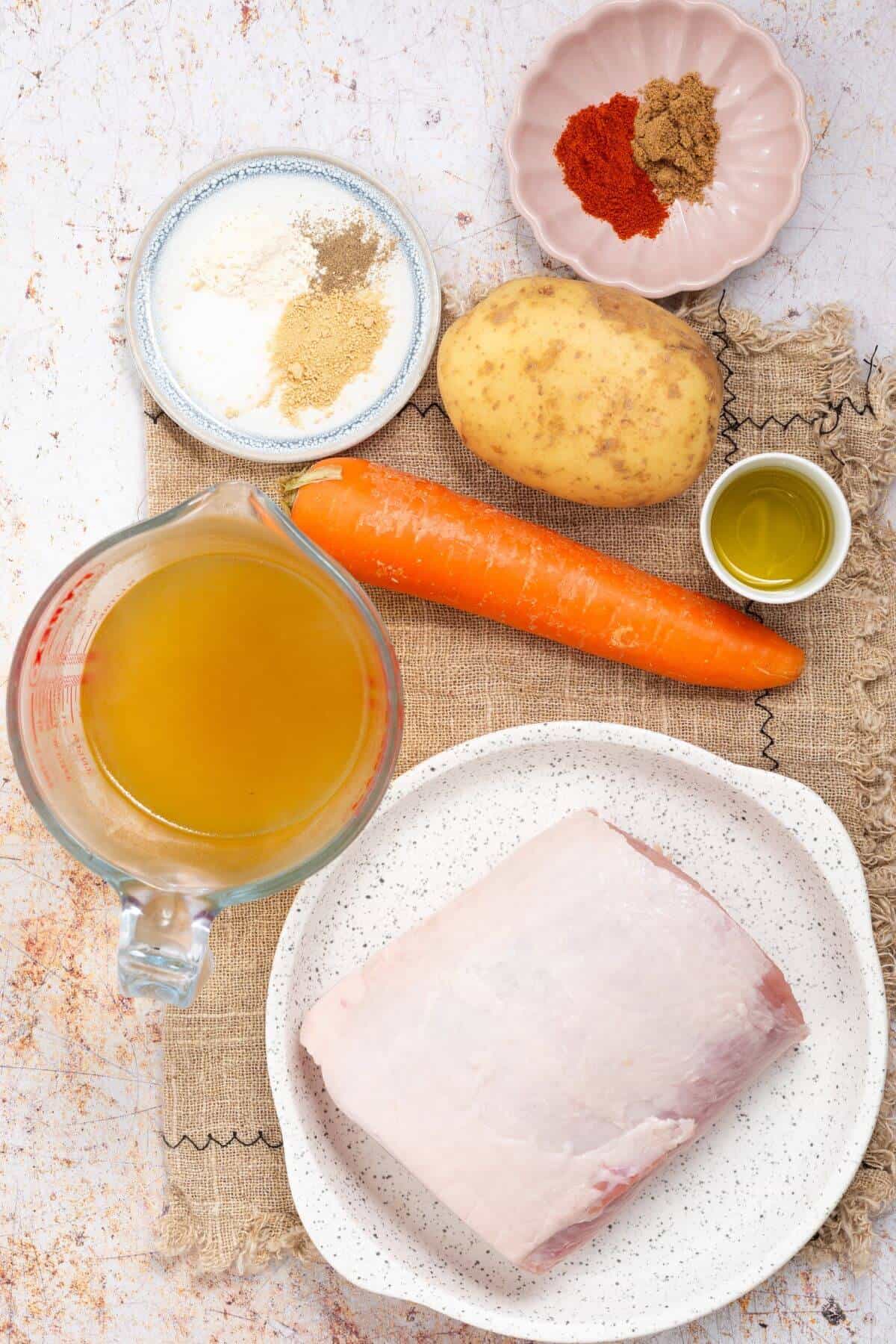 A selection of cooking ingredients: a pork roast on a plate, a carrot, a potato, a jug of broth, oil in a small dish, and various seasonings in small bowls.