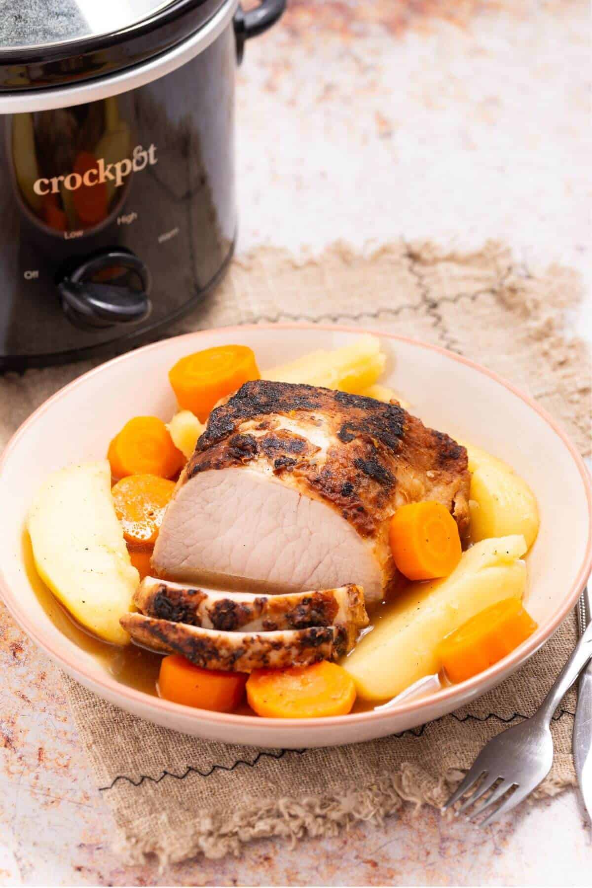 Plate with sliced roast pork, carrots, and potatoes in front of a slow cooker.