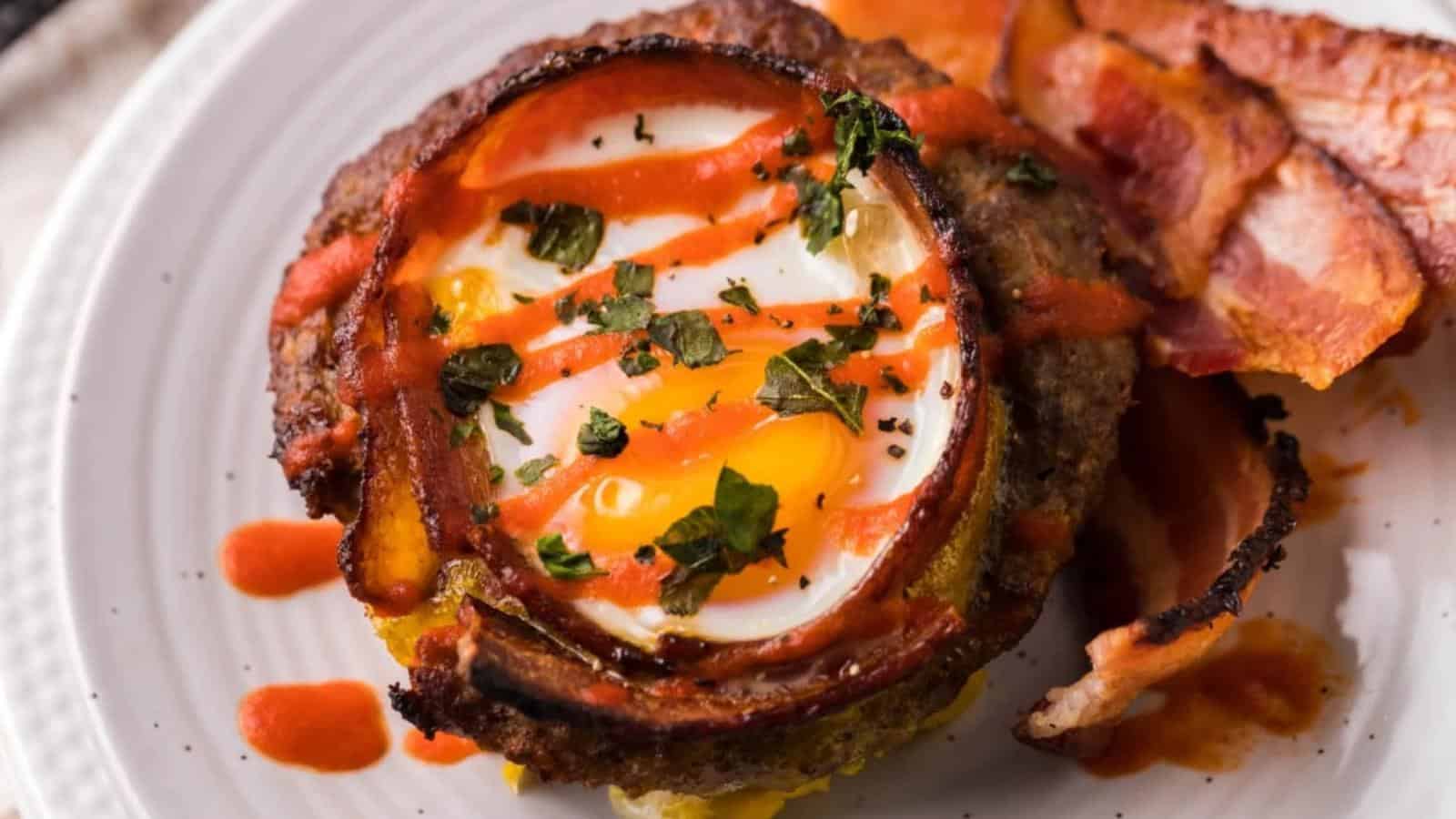 A plate with a cooked egg inside a ground meat ring, topped with herbs and drizzled with sauce, accompanied by bacon.