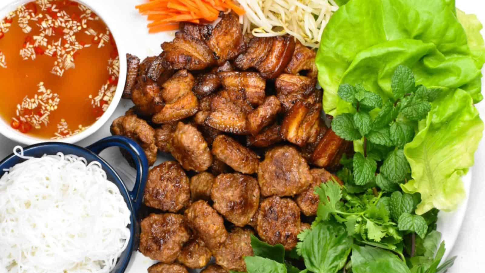 A plate with grilled meat, fresh lettuce, mint, cilantro, and a bowl of vermicelli noodles. Carrot strips and bean sprouts are beside a bowl of dipping sauce with chili and garlic.