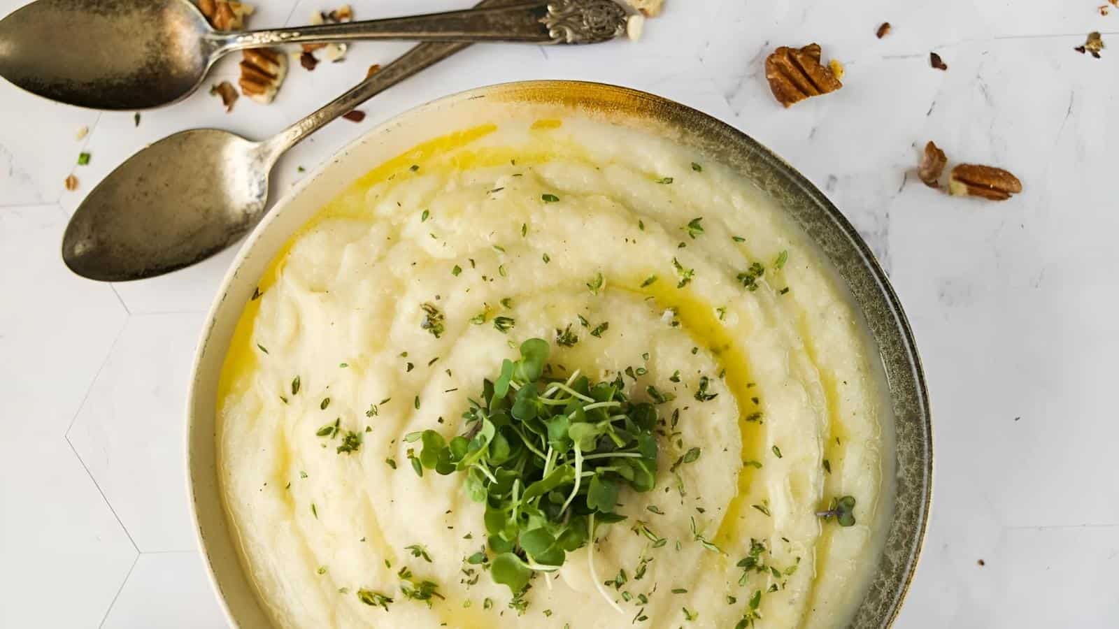 Bowl of creamy mashed potatoes garnished with green herbs, drizzled with oil. Two spoons and scattered pecans in the background on a white surface.