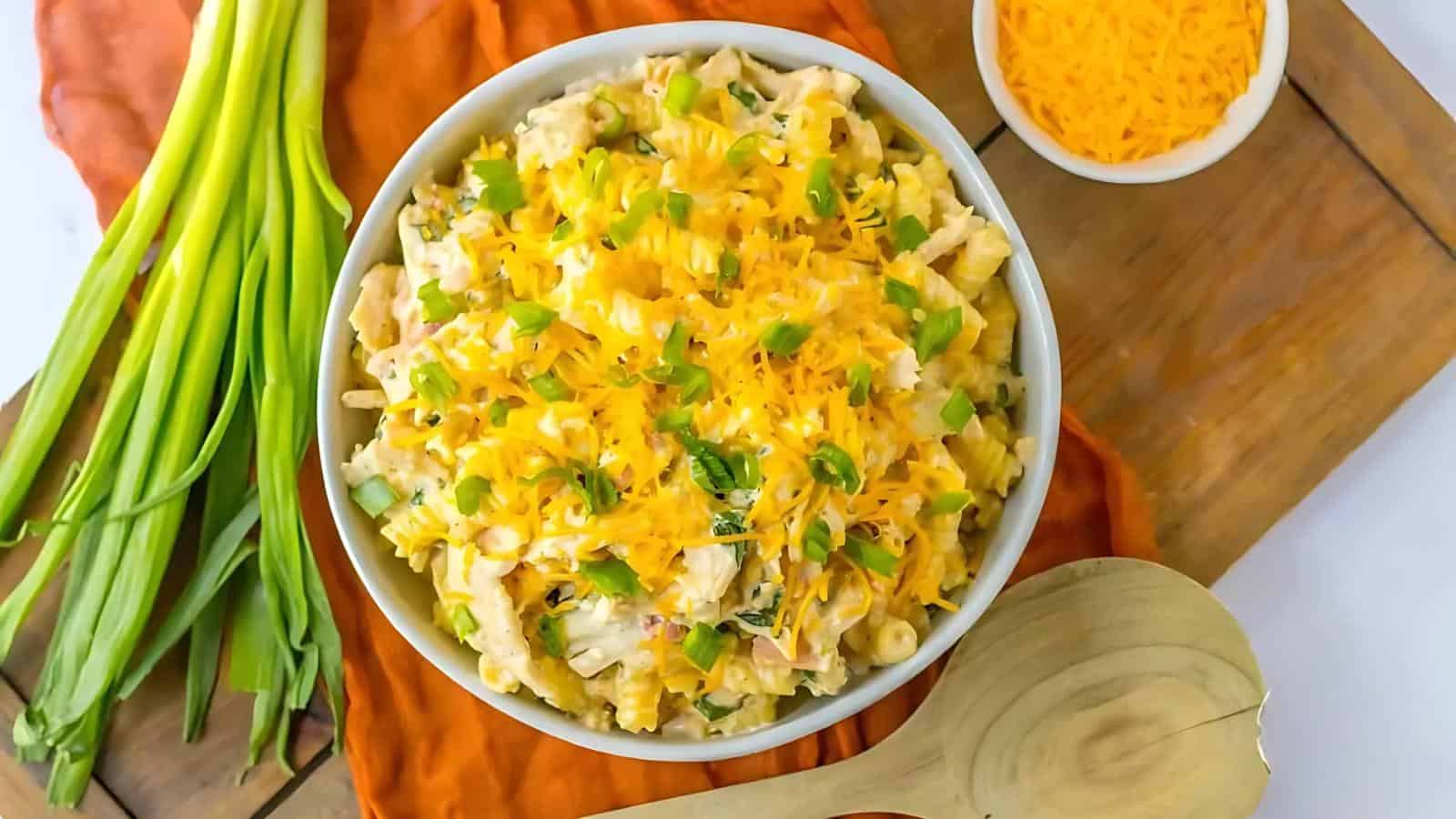 A bowl of macaroni salad topped with shredded cheese and chopped green onions. Green onions are beside the bowl. A wooden spoon and a small dish of cheese are also on the table.