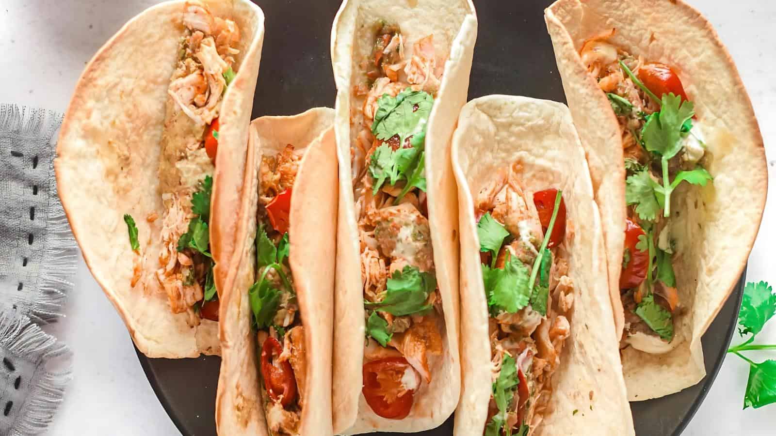 Four tacos filled with shredded chicken, cilantro, tomato, and a creamy sauce on a black plate.