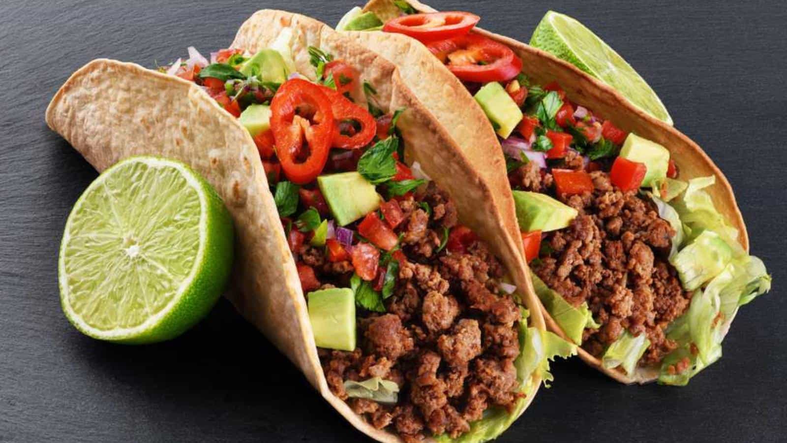 Three beef tacos with lettuce, avocado, tomatoes, and sliced red chili peppers on tortillas, accompanied by a halved lime on a dark surface.