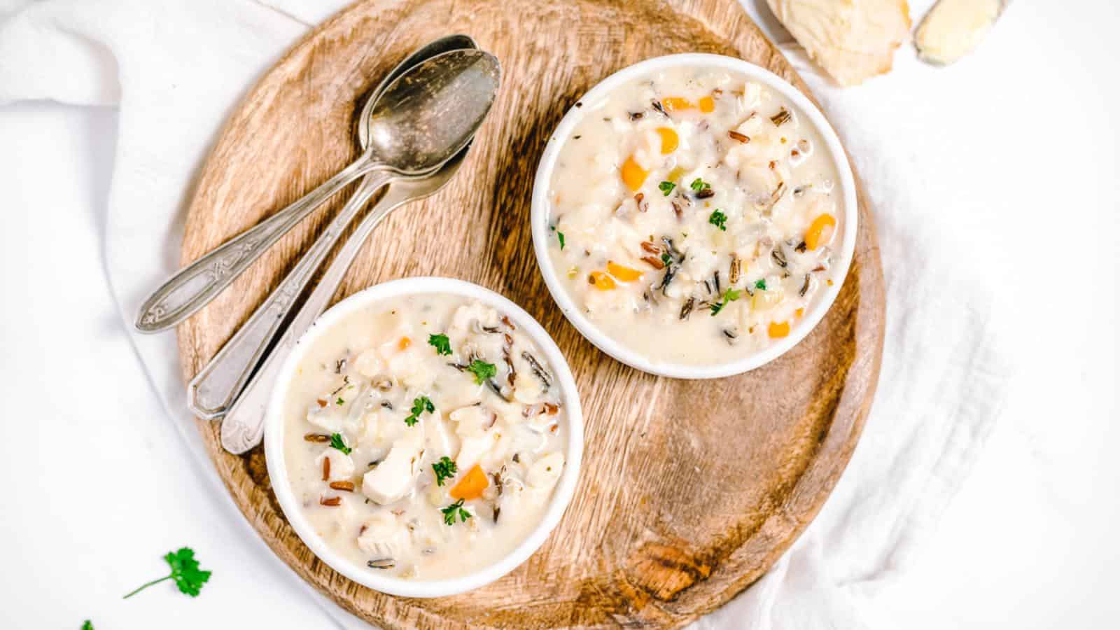 Two bowls of creamy chicken and wild rice soup on a round wooden board with spoons beside them.