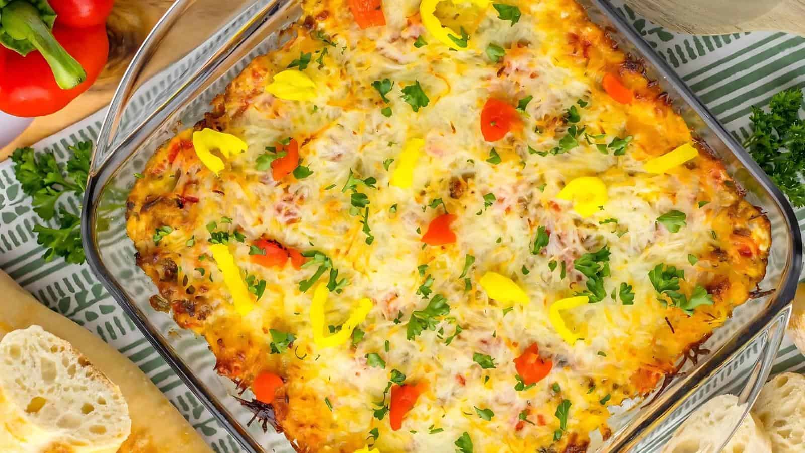Baked casserole topped with melted cheese, yellow and red bell peppers, and fresh parsley, served in a glass dish with slices of bread nearby.