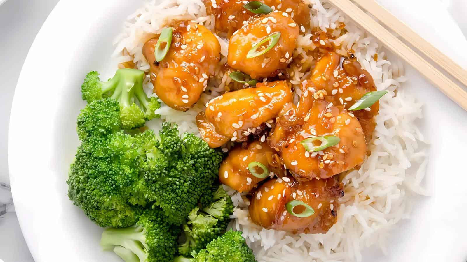 A white plate with white rice topped with sesame chicken and green onions, accompanied by steamed broccoli.