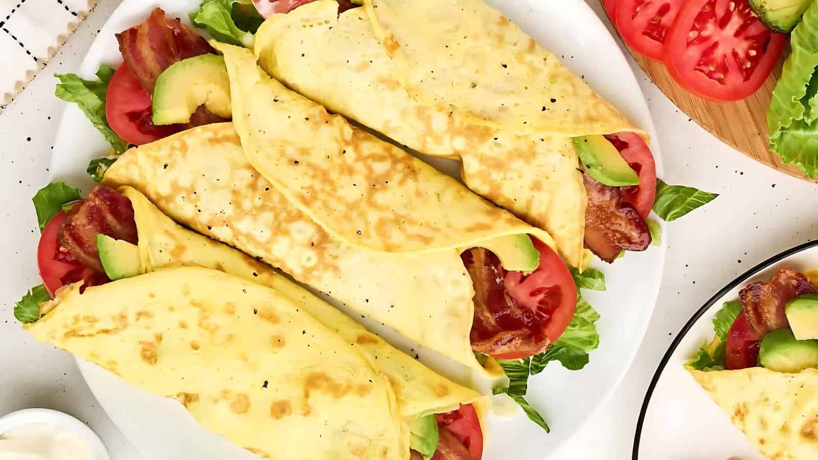 Crepes filled with bacon, lettuce, avocado, and tomato are arranged on a white plate. Sliced tomatoes and lettuce are visible on the side.