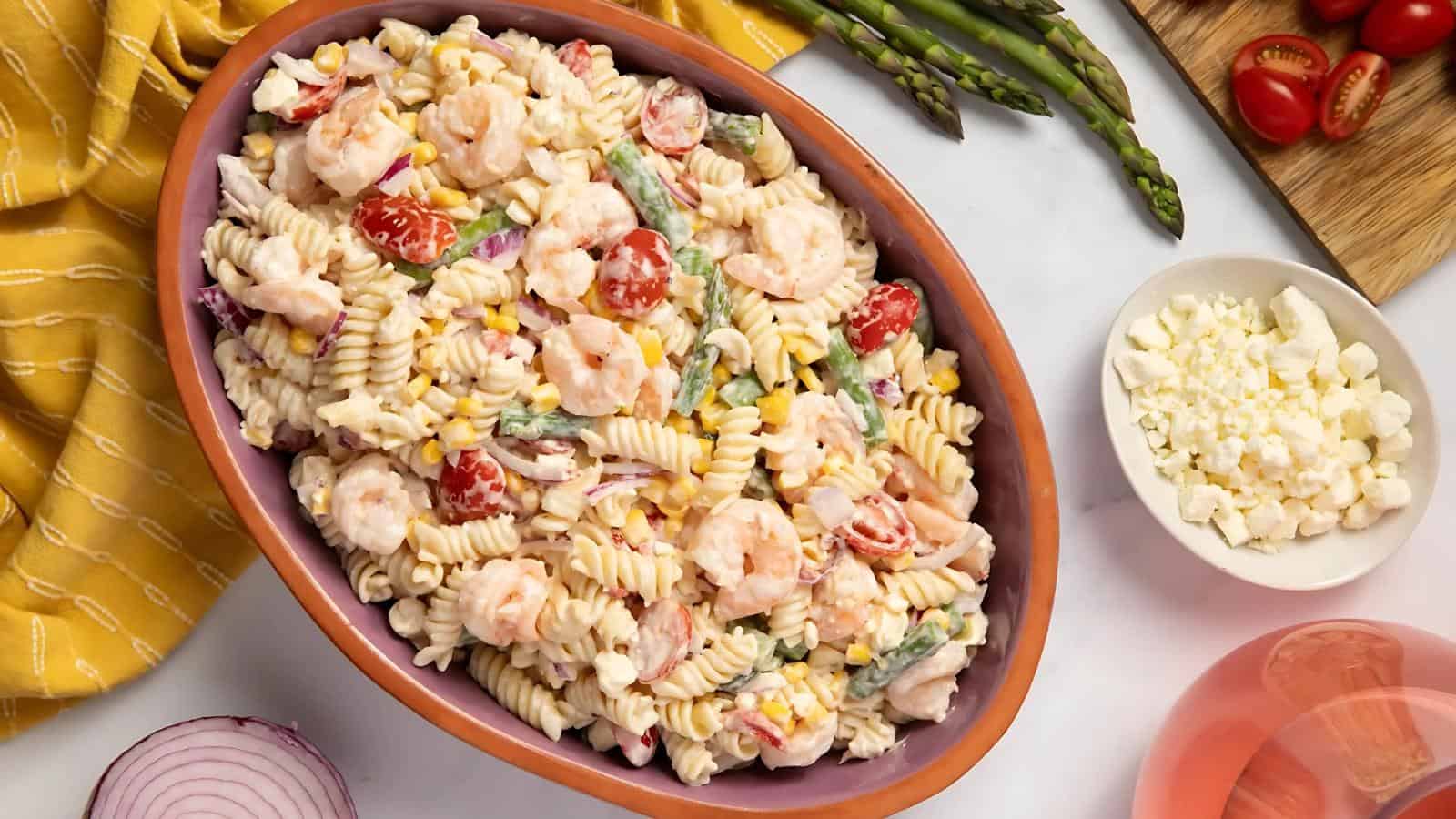 A bowl of creamy pasta salad with shrimp, cherry tomatoes, corn, and asparagus, surrounded by ingredients like cherry tomatoes, asparagus, diced cheese, and a yellow napkin.