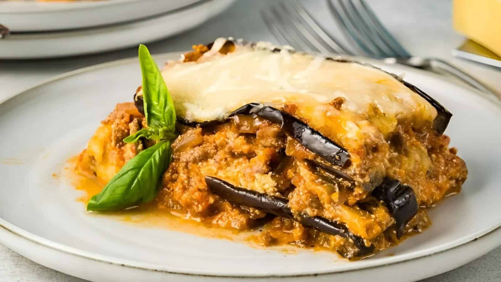 A slice of eggplant lasagna with cheese and meat sauce is served on a white plate, garnished with a basil leaf.
