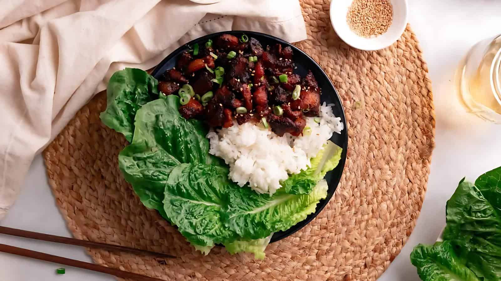 A black plate with rice, stir-fried meat, and chopped green onions is served with lettuce on the side. Chopsticks and a cloth napkin are on a woven placemat.