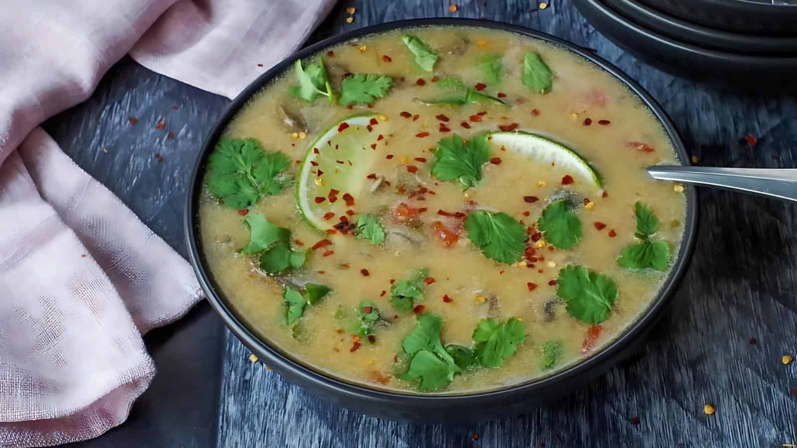 Bowl of soup garnished with lime slices, cilantro, and red chili flakes, placed on a dark surface with a pink cloth nearby.