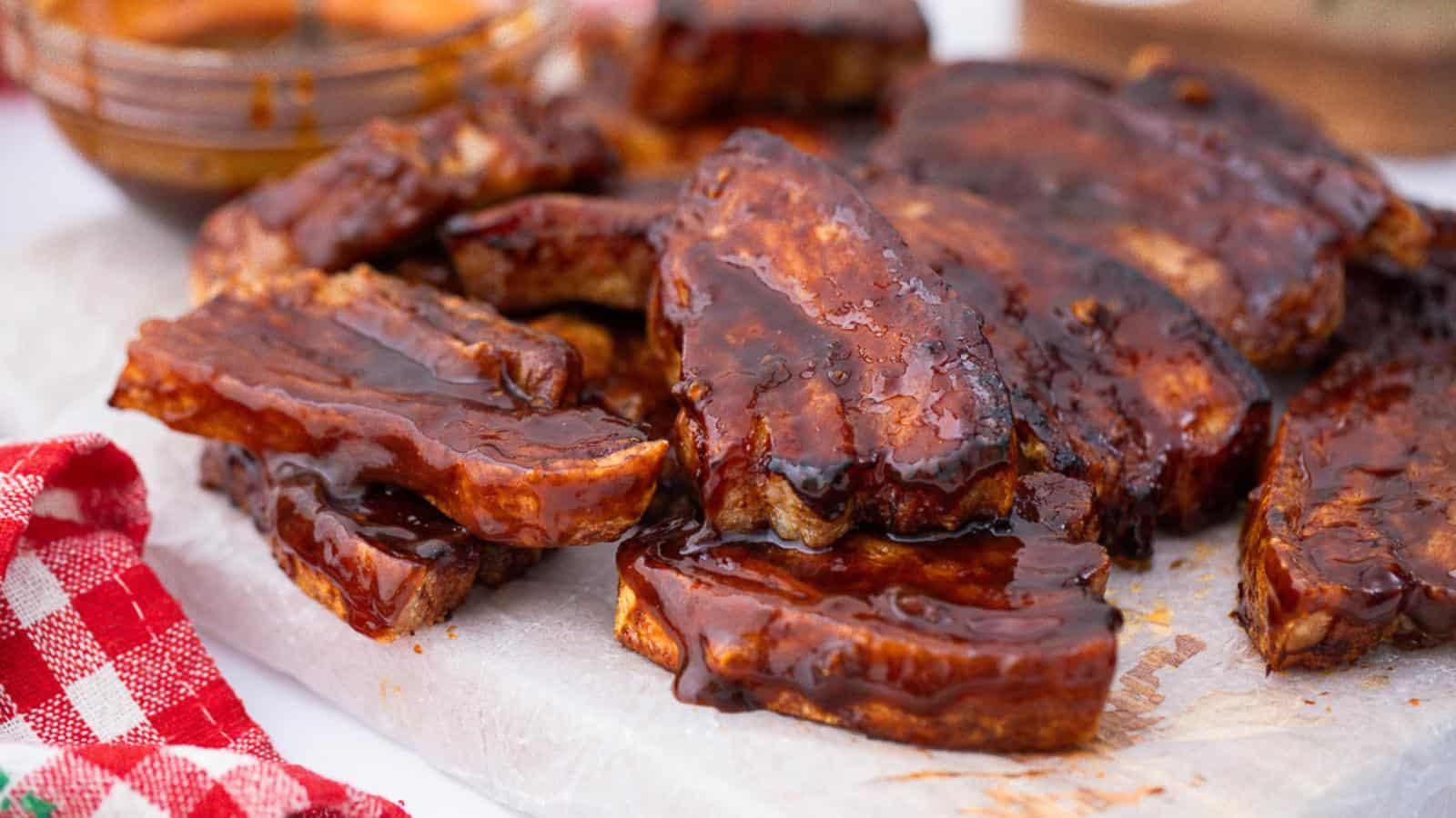 Barbecue ribs covered in a glossy sauce are stacked on a paper-lined board. A checkered cloth is visible in the corner.