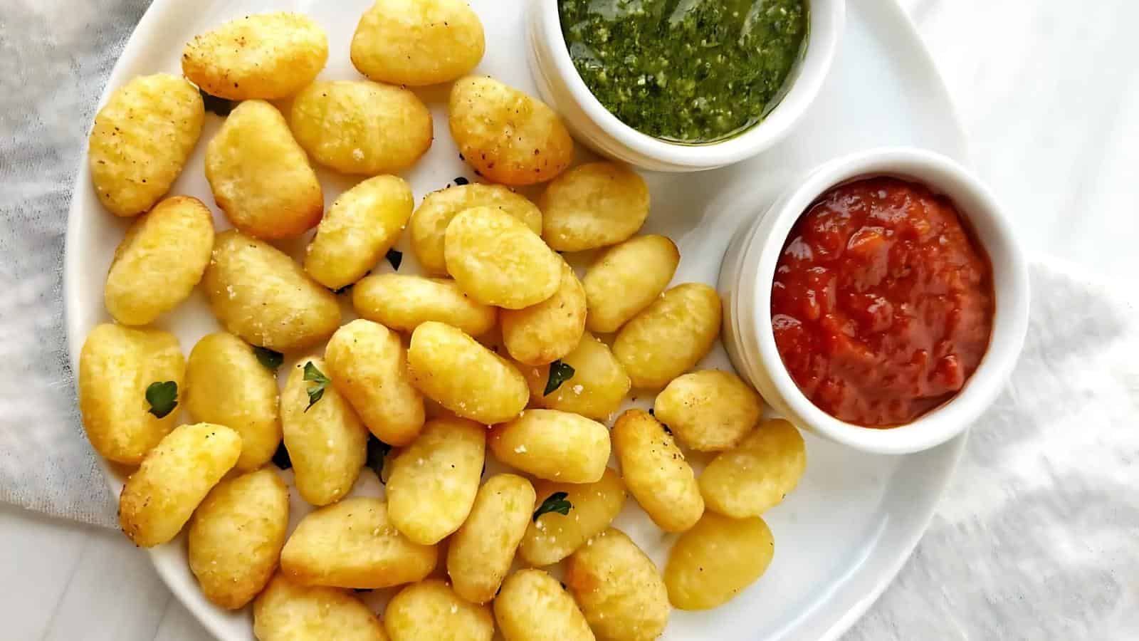 A plate of crispy potato gnocchi with dipping sauces, one green pesto and one red tomato-based, on a white plate.