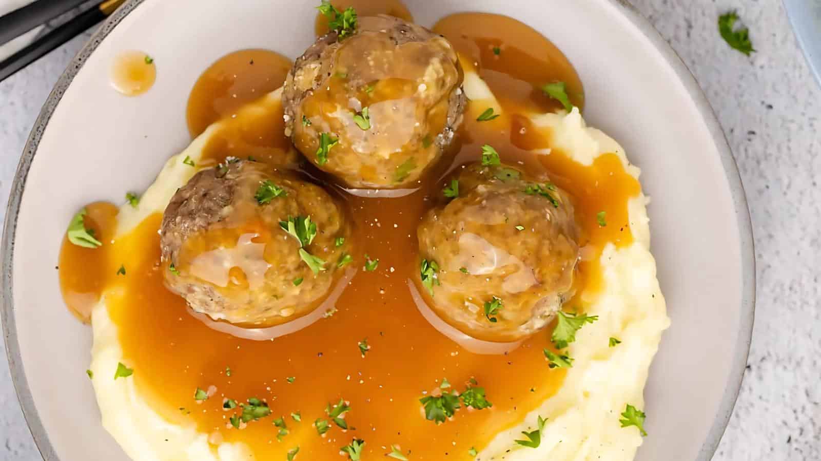 A plate of mashed potatoes topped with three meatballs, covered in gravy, and garnished with parsley.