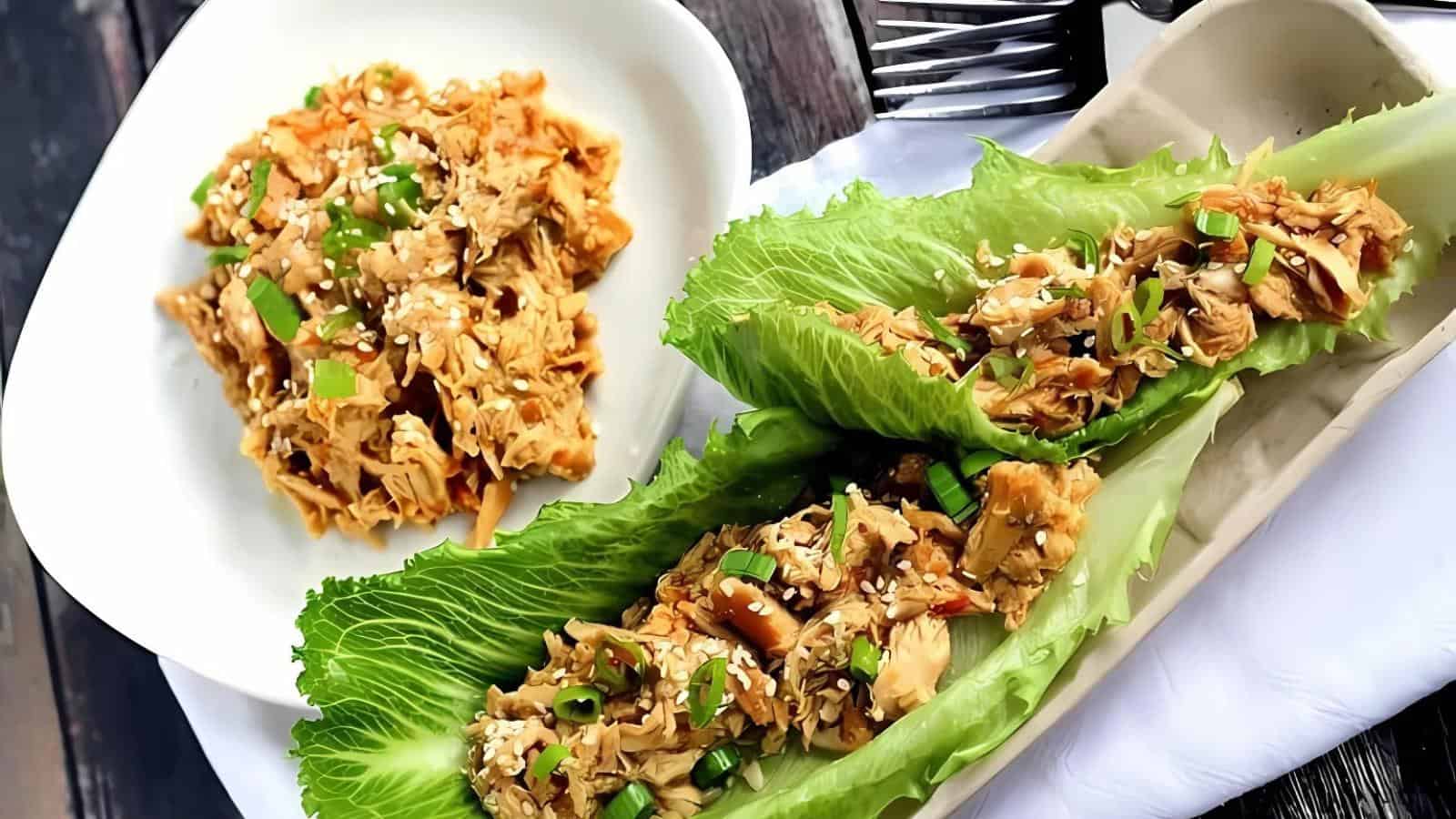 Shredded chicken with sesame seeds and green onions served on lettuce leaves and in a bowl, placed on a white napkin with a fork beside it.