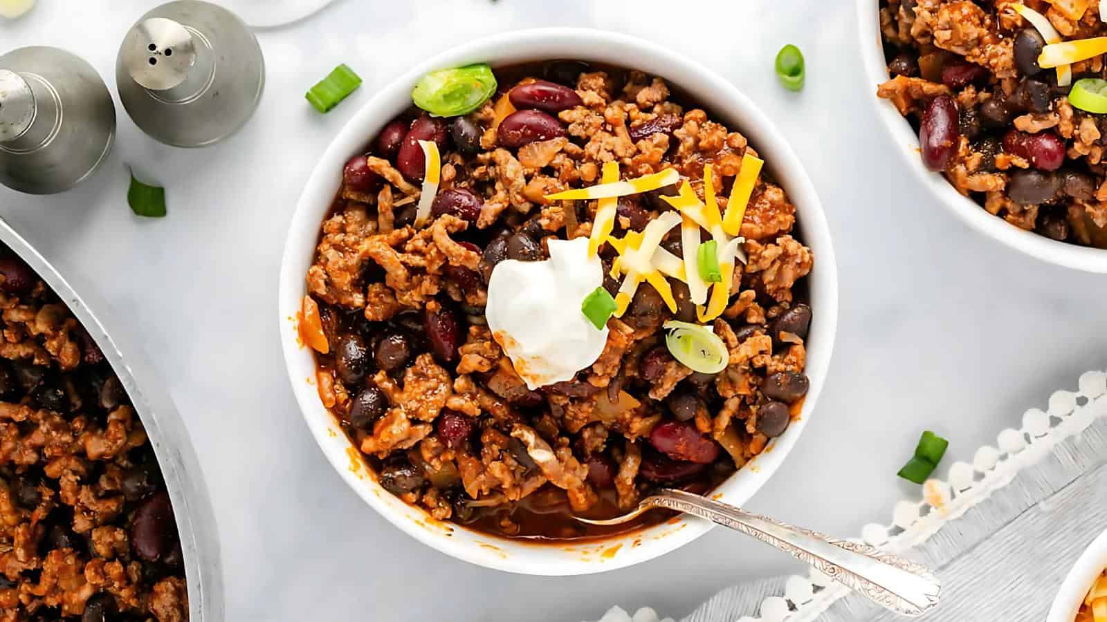 Bowl of chili with ground meat, beans, sour cream, shredded cheese, and green onions, accompanied by a spoon.