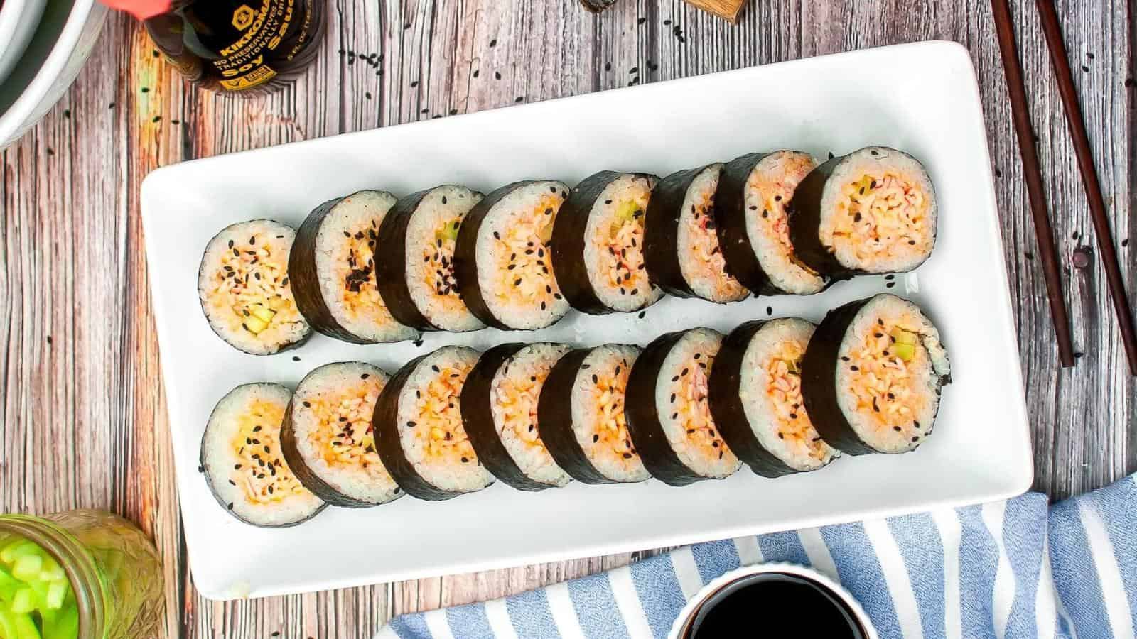 A rectangular plate with neatly arranged sliced sushi rolls, filled with rice and topped with sesame seeds. Chopsticks, soy sauce, and a striped cloth are nearby on a wooden surface.