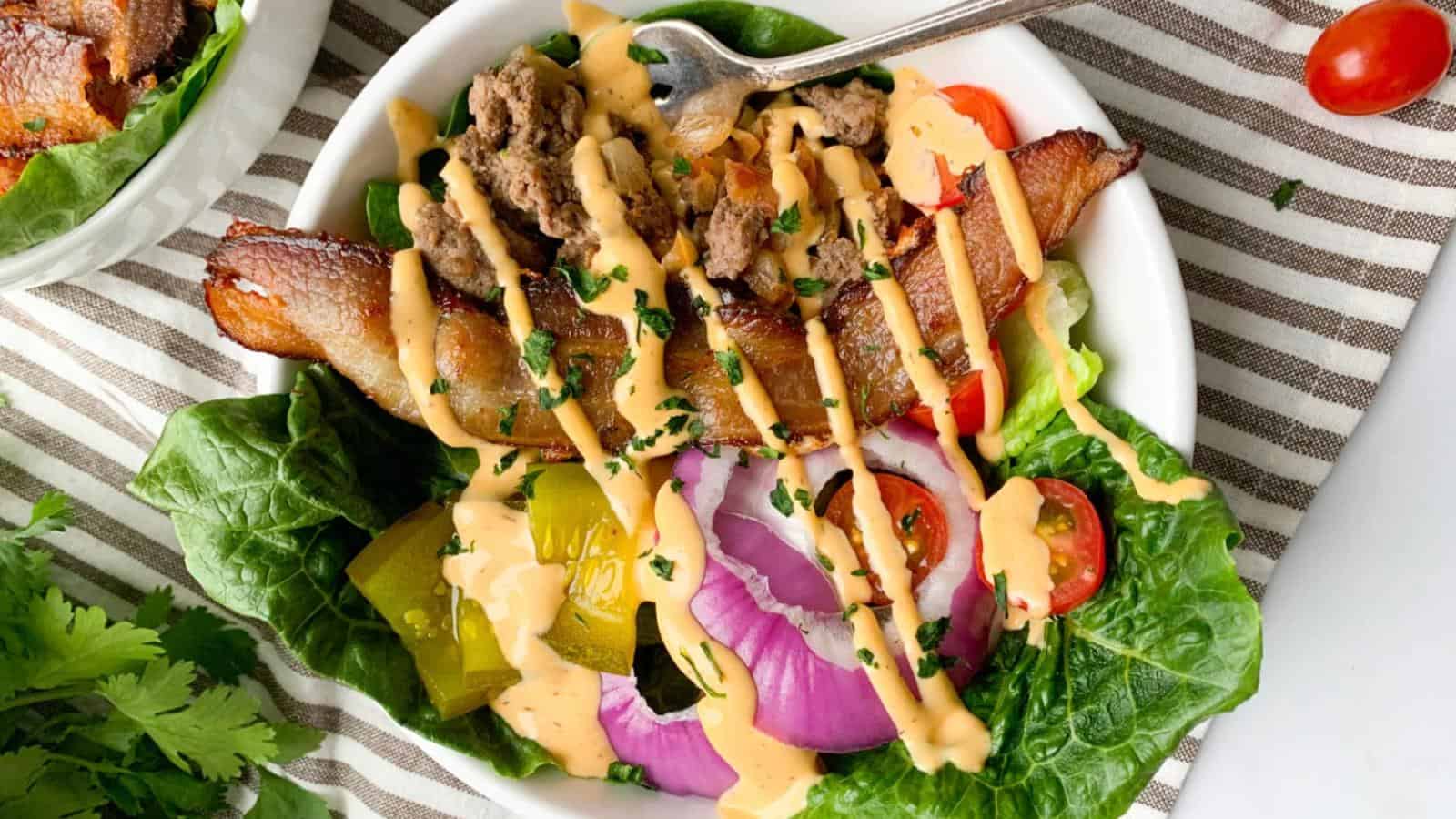 Salad bowl with bacon, ground meat, lettuce, red onion, tomatoes, pickles, and drizzled sauce on a striped cloth.