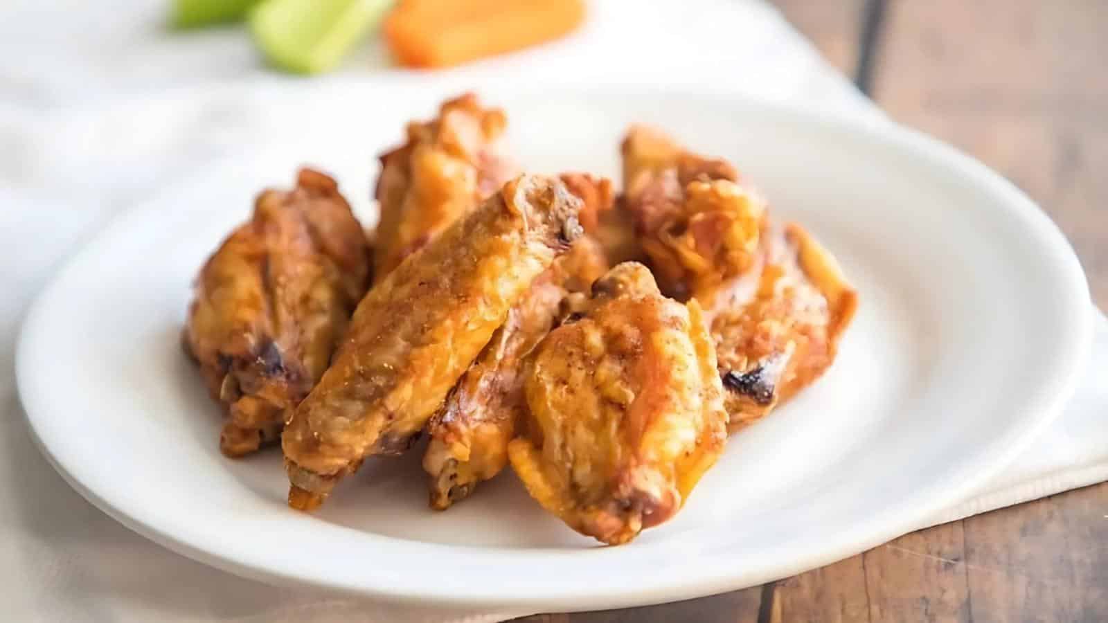 A plate of eight cooked chicken wings on a white plate with blurred celery and carrot sticks in the background.