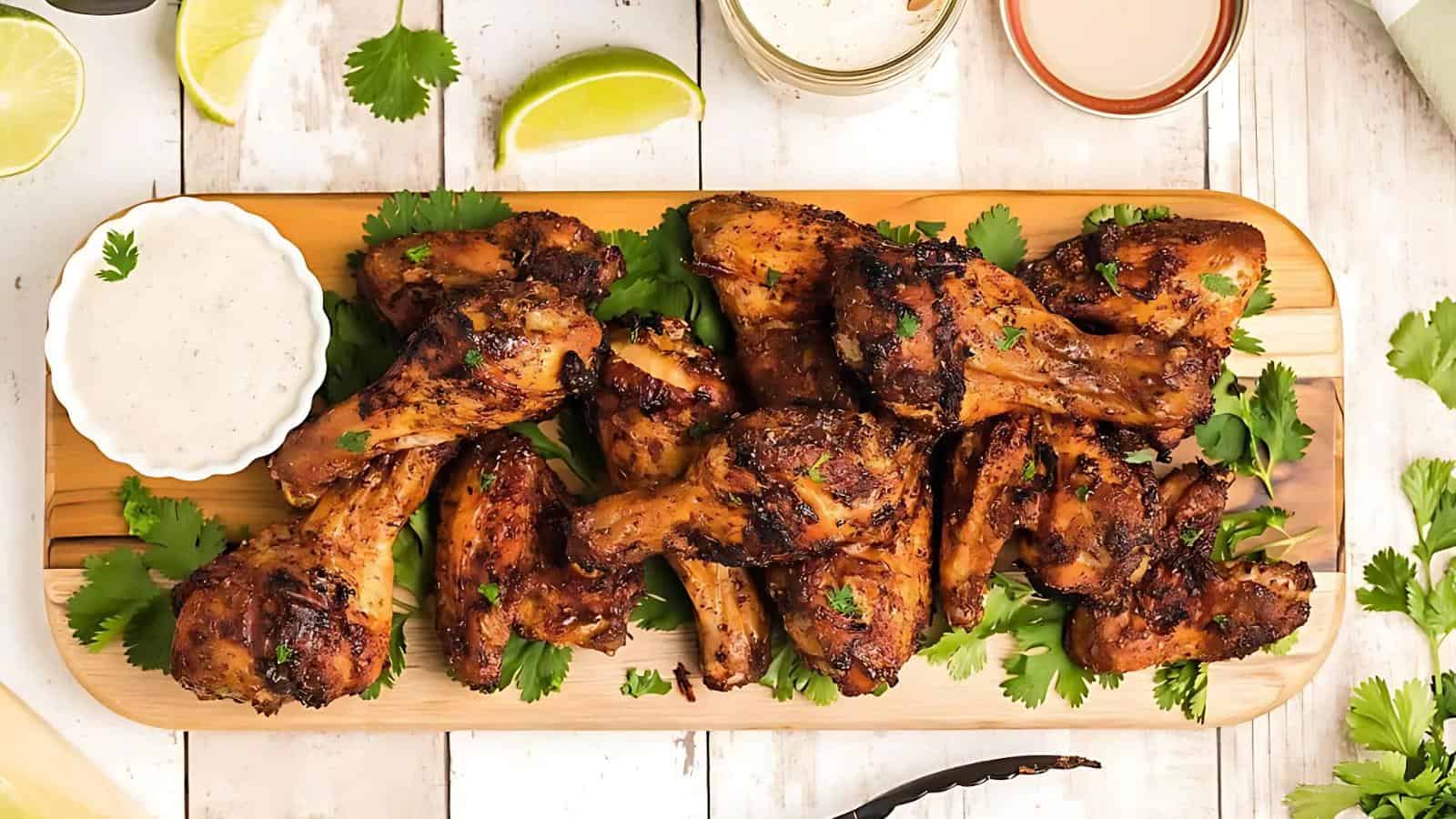 A wooden tray with grilled chicken wings, garnished with cilantro. Accompanied by a small bowl of dipping sauce and lime wedges on a white wooden surface.