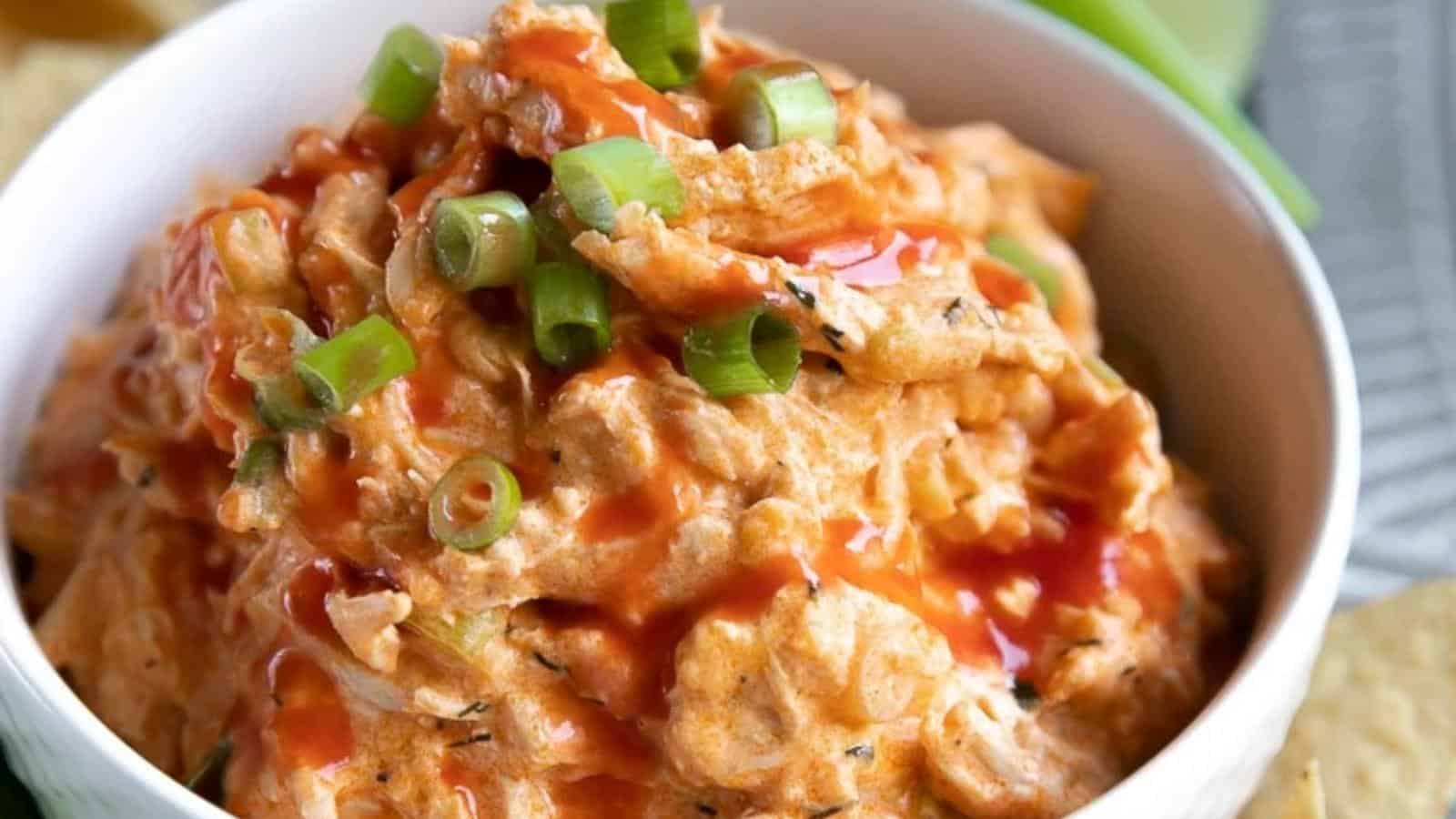 A bowl of buffalo chicken dip topped with chopped green onions and drizzled with hot sauce.