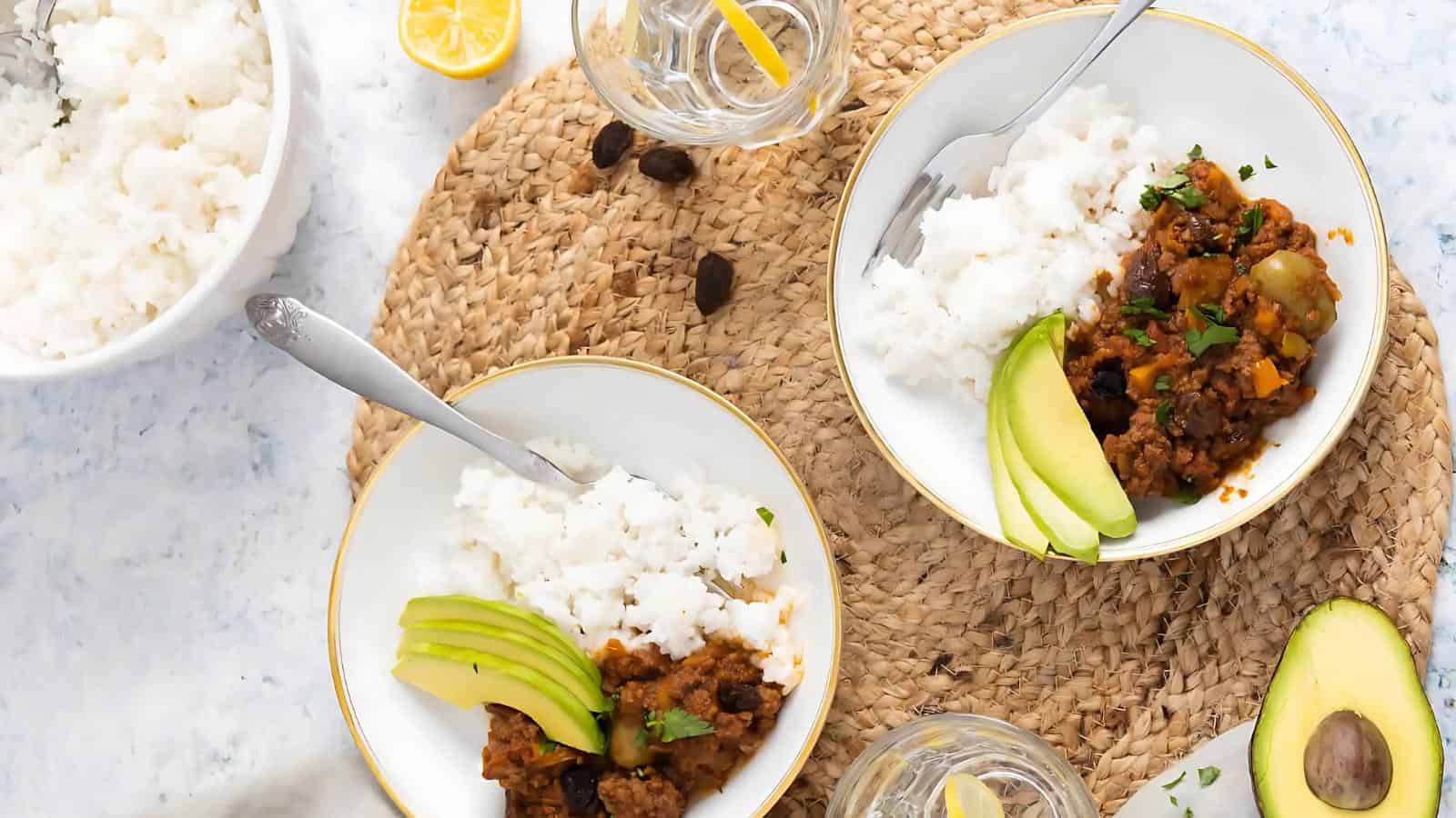 Two plates of rice topped with avocado slices and a vegetable stew, placed on a woven mat. A bowl of white rice, lemon slices, and glasses of water are nearby.