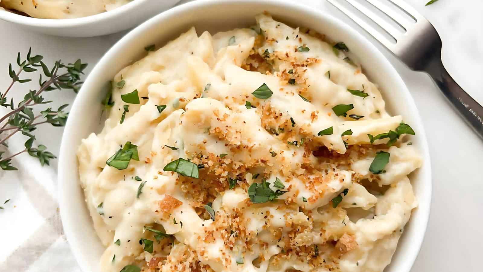 A bowl of creamy pasta topped with breadcrumbs and parsley, with a fork and fresh herbs on the side.