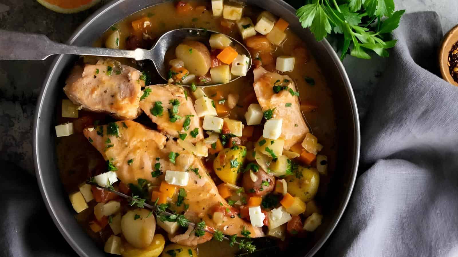Bowl of salmon stew with vegetables, herbs, and diced cheese, garnished with parsley, with a spoon in the dish.