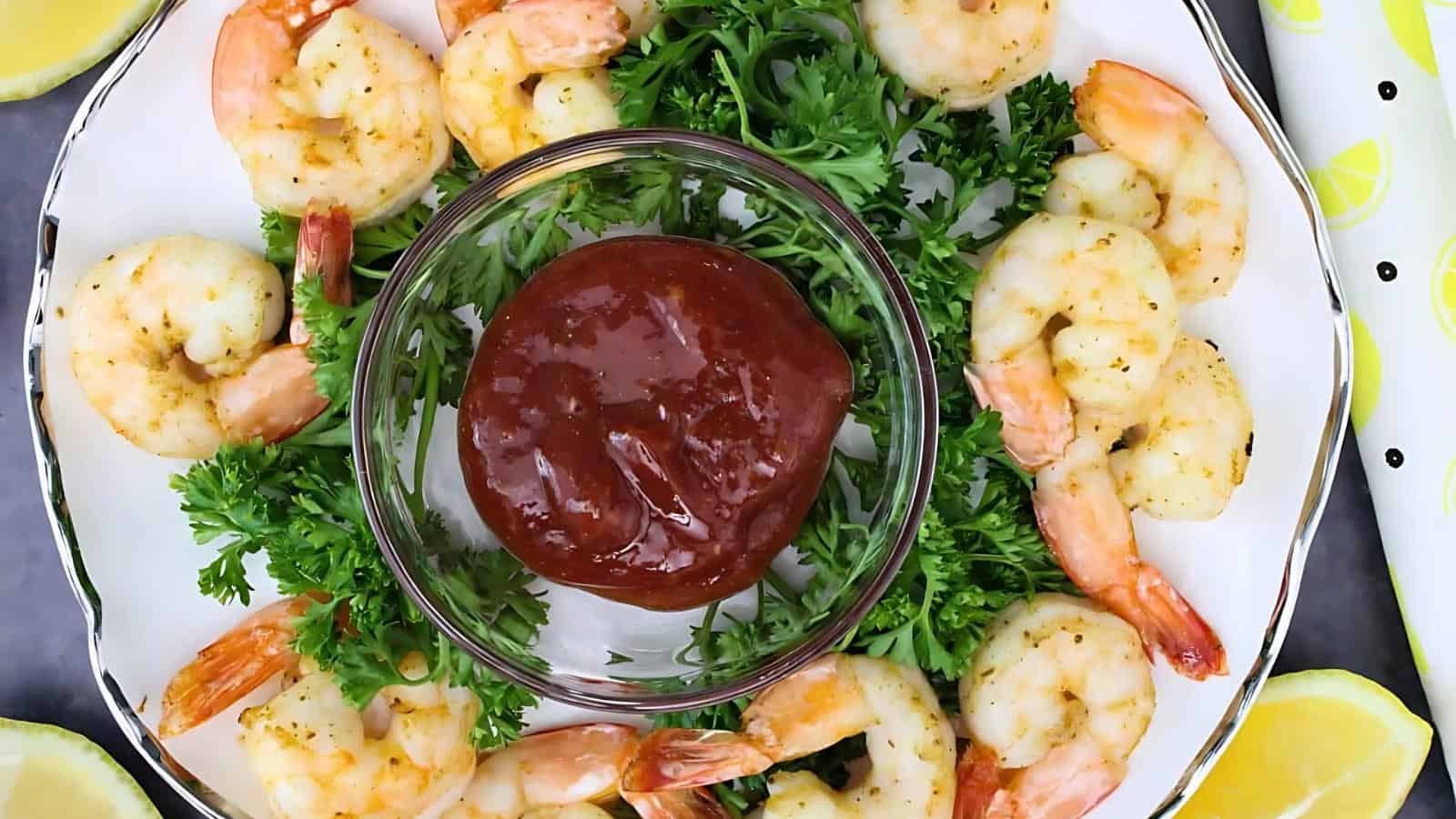 Cooked shrimp arranged in a circle on a plate with parsley, surrounding a central bowl of cocktail sauce.