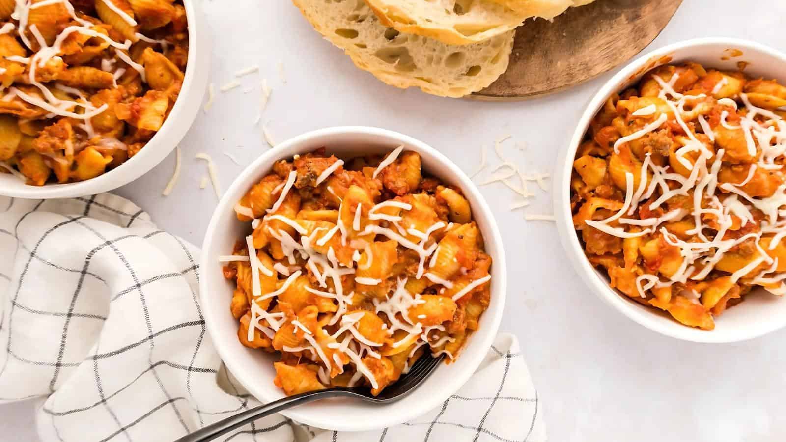 Three bowls of pasta with tomato sauce and shredded cheese, accompanied by sliced bread and a checkered napkin.
