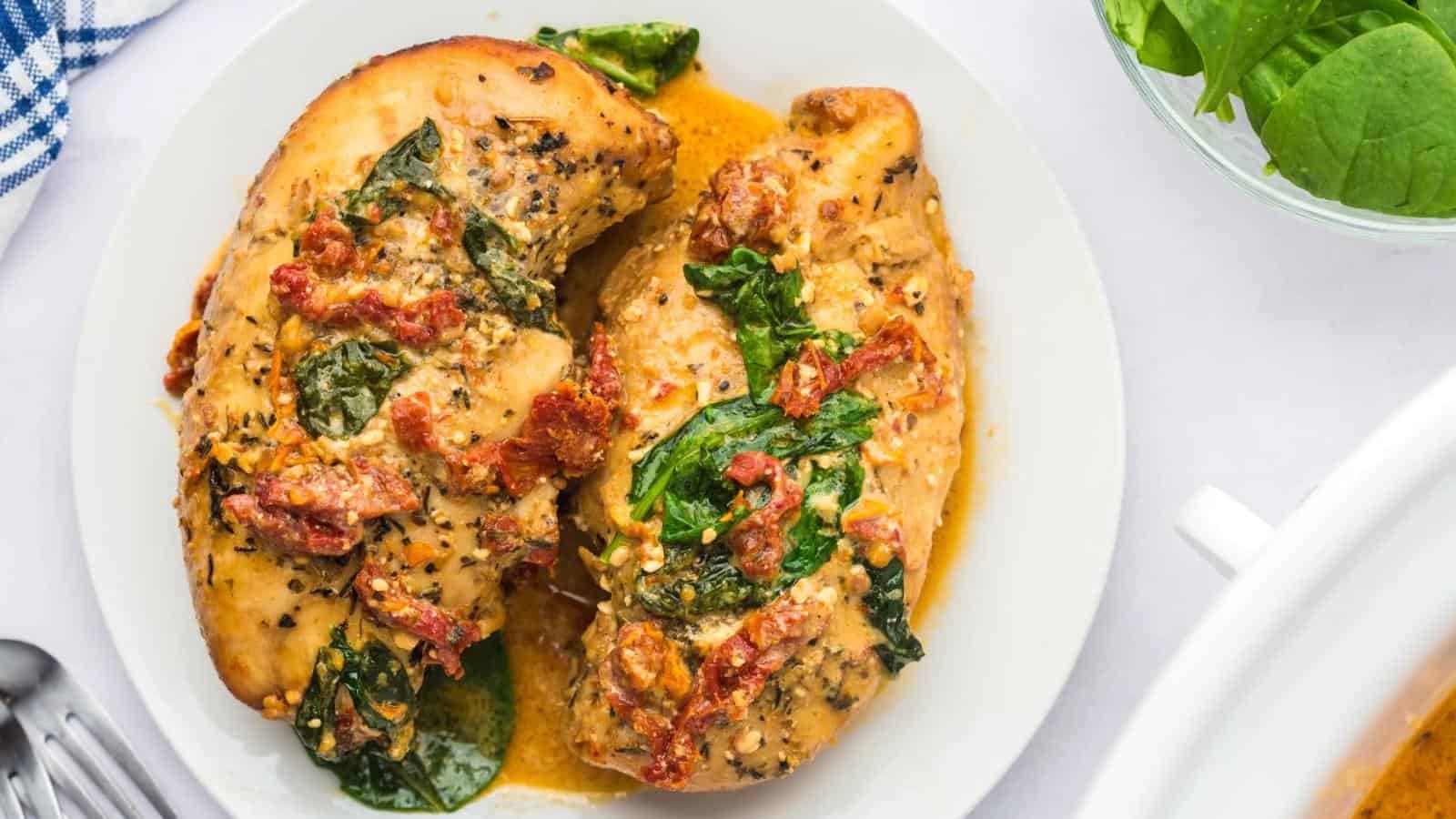 Two cooked chicken breasts on a white plate, topped with spinach and sun-dried tomatoes in a creamy sauce.