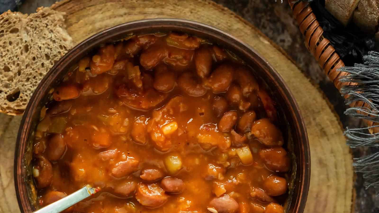A bowl of baked beans with tomato sauce is placed on a wooden surface. A slice of brown bread is visible beside the bowl.