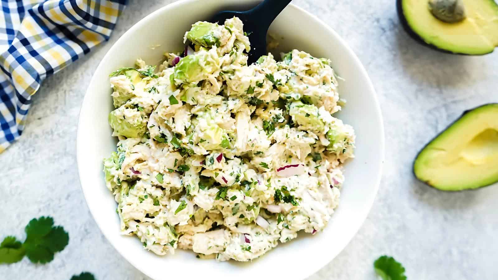 A bowl of chicken salad with avocado, red onion, and herbs, with a spoon. An avocado half and cilantro leaves are nearby on a white surface.
