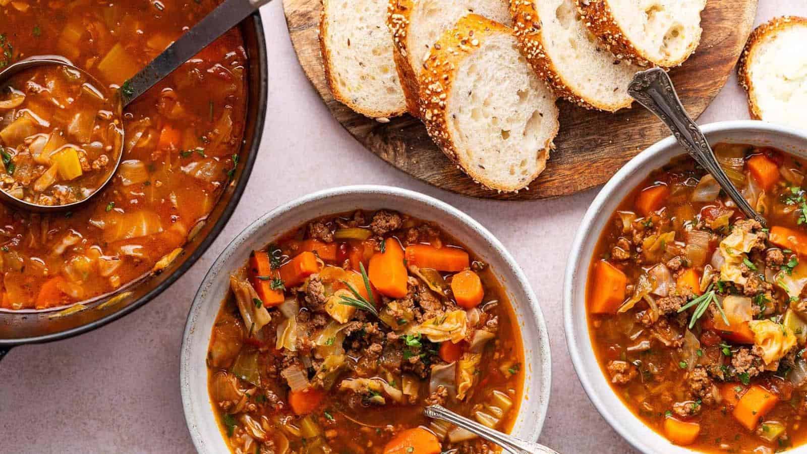 Two bowls of vegetable and meat soup with spoons, a pot with a ladle, and slices of sesame bread on a wooden board.