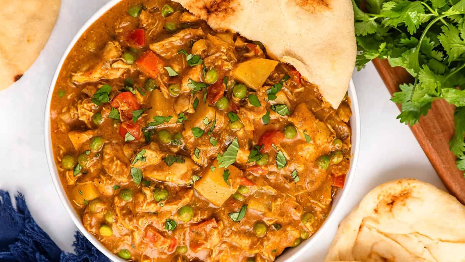 A bowl of curry with peas, potatoes, and tomatoes, garnished with cilantro, served with naan bread on a white surface.