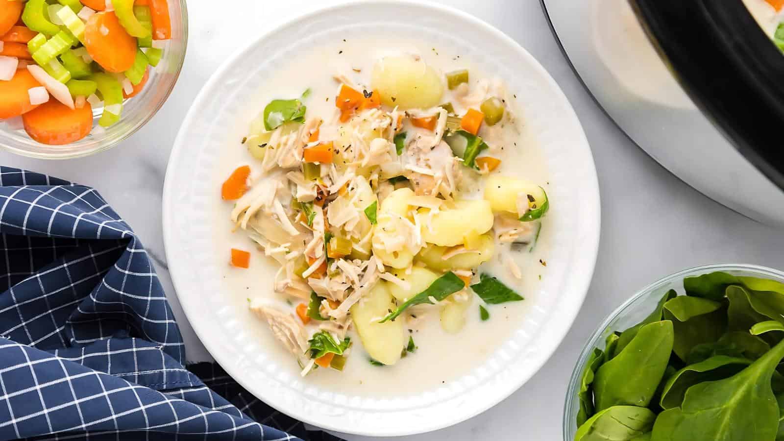 A white plate with creamy chicken soup containing pieces of chicken, gnocchi, carrots, celery, and spinach. Nearby are bowls of chopped vegetables and fresh spinach. A blue checkered cloth is on the side.