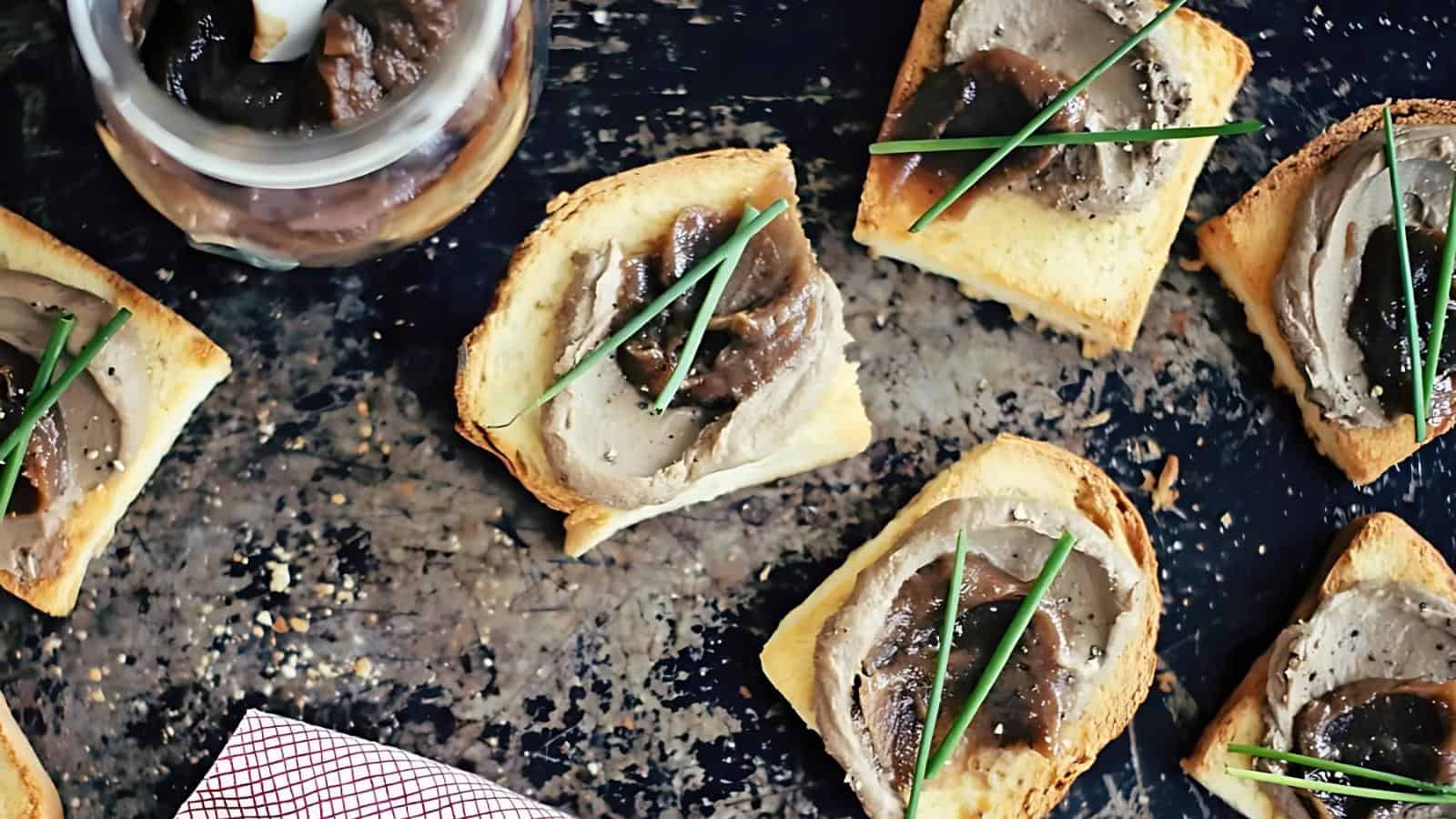 Top view of toasted bread slices topped with pâté, caramelized onions, and chives on a dark textured surface.