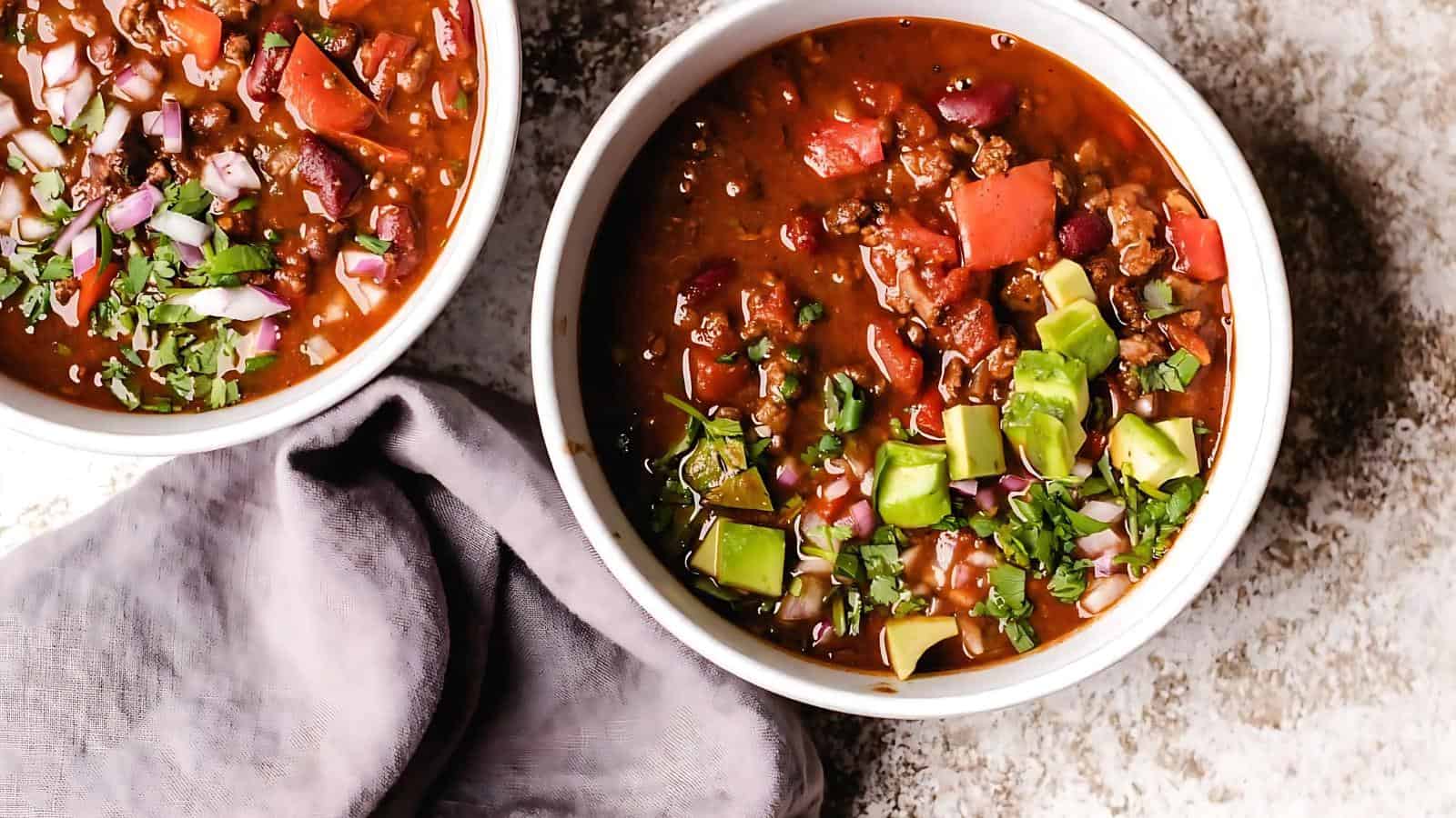 Two bowls of chili topped with avocado, red onions, and cilantro, next to a gray cloth on a textured surface.