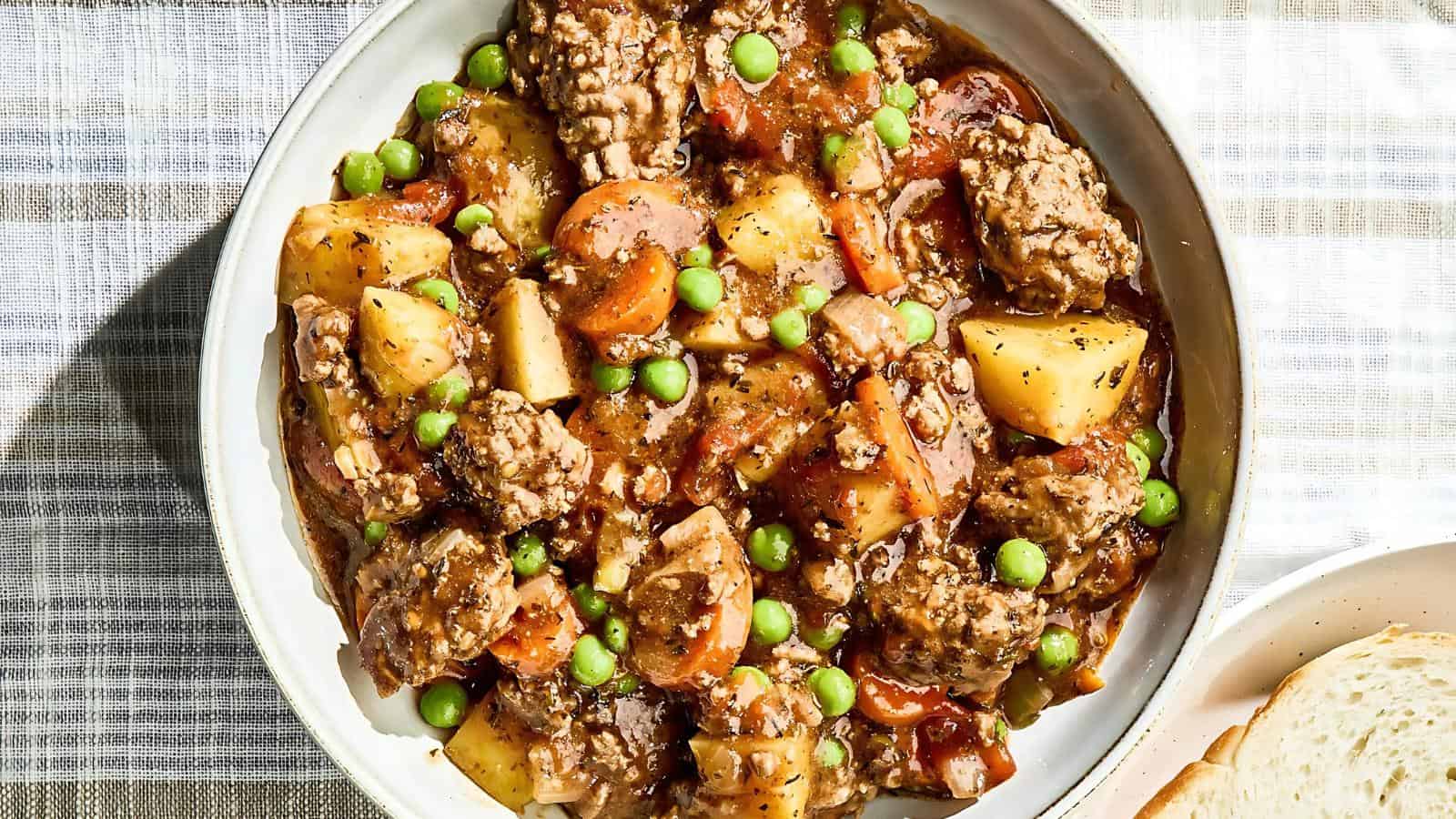 A bowl of beef stew with potatoes, carrots, peas, and a seasoned broth on a plaid tablecloth.